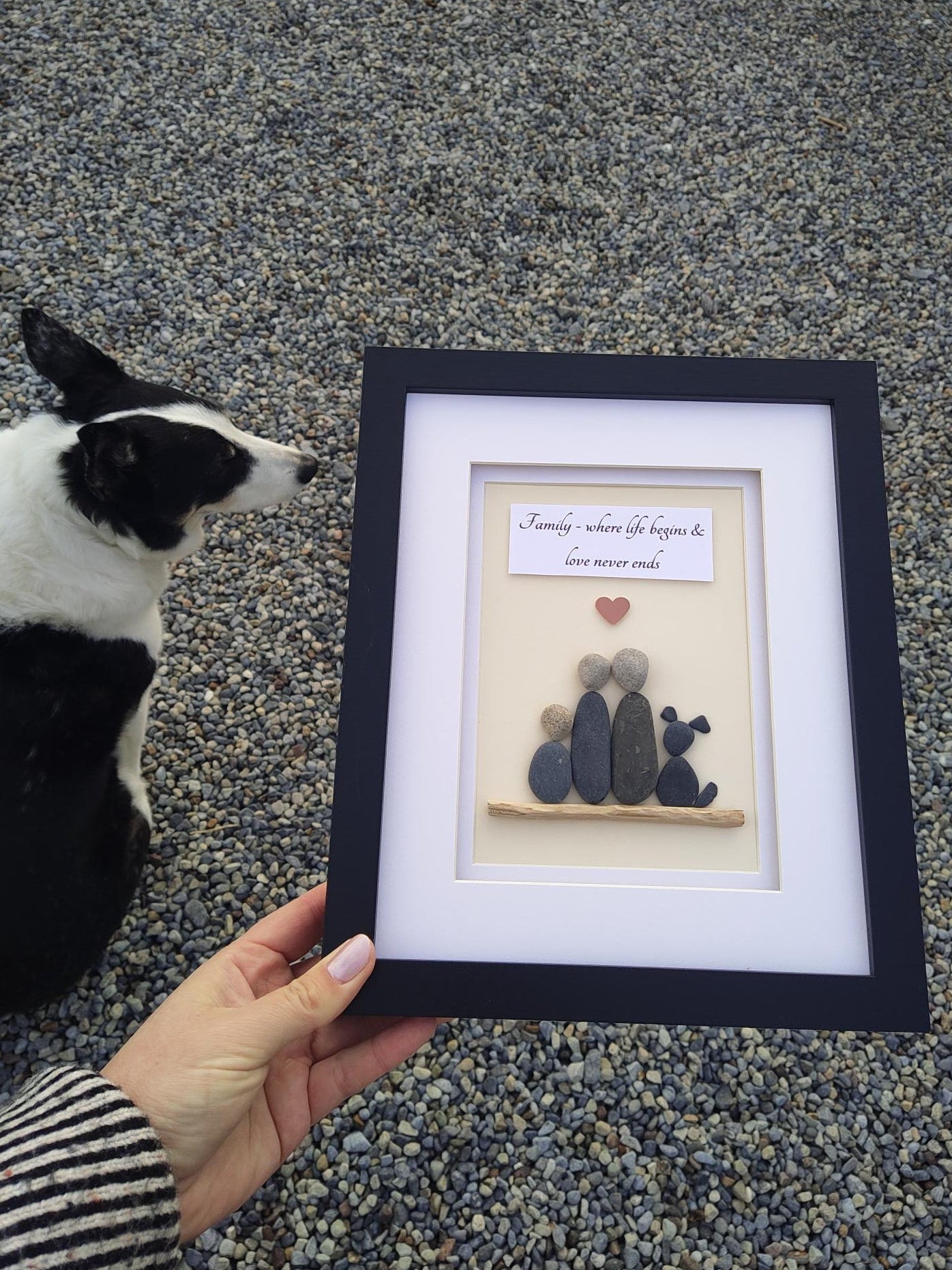 A hand holds a black-framed, white-matted custom sea-foam art piece featuring small stones and a small heart, with Jack looking up at it from below on a gravel surface.