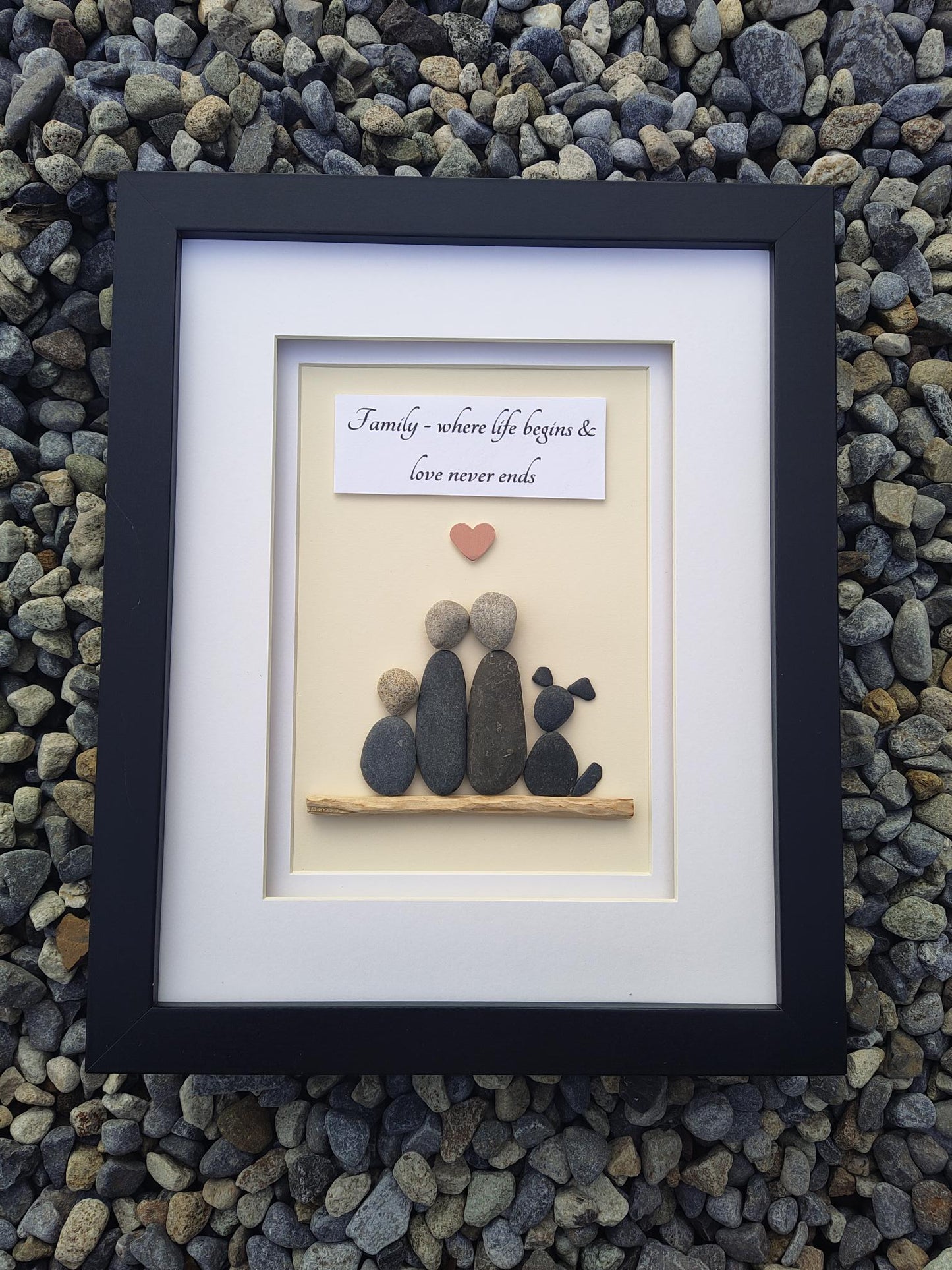 A framed piece of personalized stone art displayed on a bed of small gray pebbles. The artwork features smooth dark and light gray stones arranged to form a silhouette, a small red heart, and a white card with black script text, all set inside a white mat and a black frame.