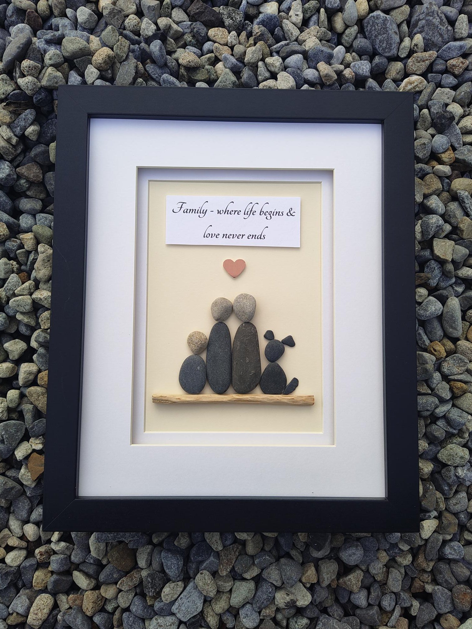 A framed piece of personalized stone art displayed on a bed of small gray pebbles. The artwork features smooth dark and light gray stones arranged to form a silhouette, a small red heart, and a white card with black script text, all set inside a white mat and a black frame.