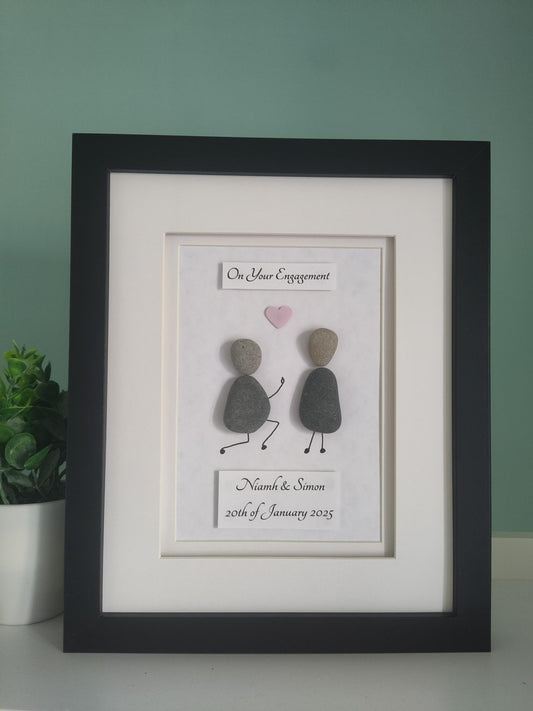 A framed pebble art artwork as a gift, with one figure on bended knee proposing to the other with a heart and the words 'On Your Engagement' displayed, along with personalized details.