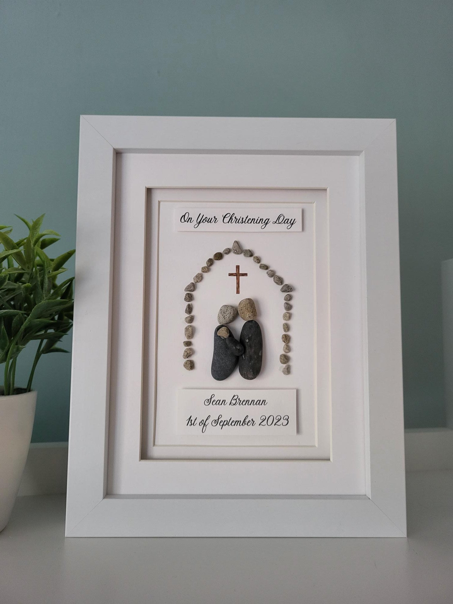 A handmade pebbleart gift featuring two parents with their baby under a cross surrounded by decorative stones, with a white frame. It has text that reads 'On Your Christening Day' with the babys name and Christening date