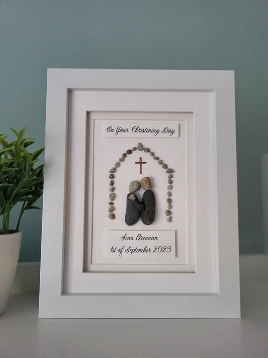 A handmade pebbleart gift featuring two parents with their baby under a cross surrounded by decorative stones, with a white frame. It has text that reads 'On Your Christening Day' with the babys name and Christening date