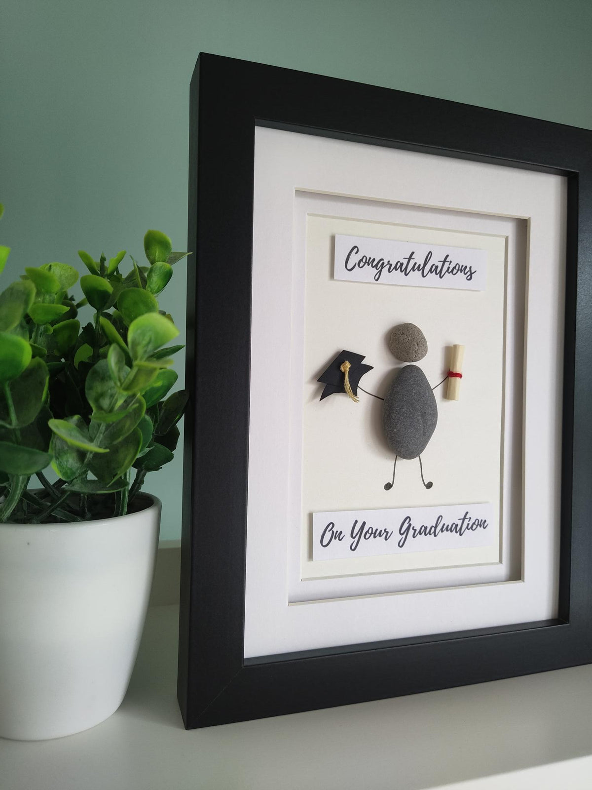 A side angle of a framed pebble art gift with the title 'CONGRATULATIONS ON YOUR GRADUATION' displayed, alongside a representation of a graduation cap and a graduation scroll.