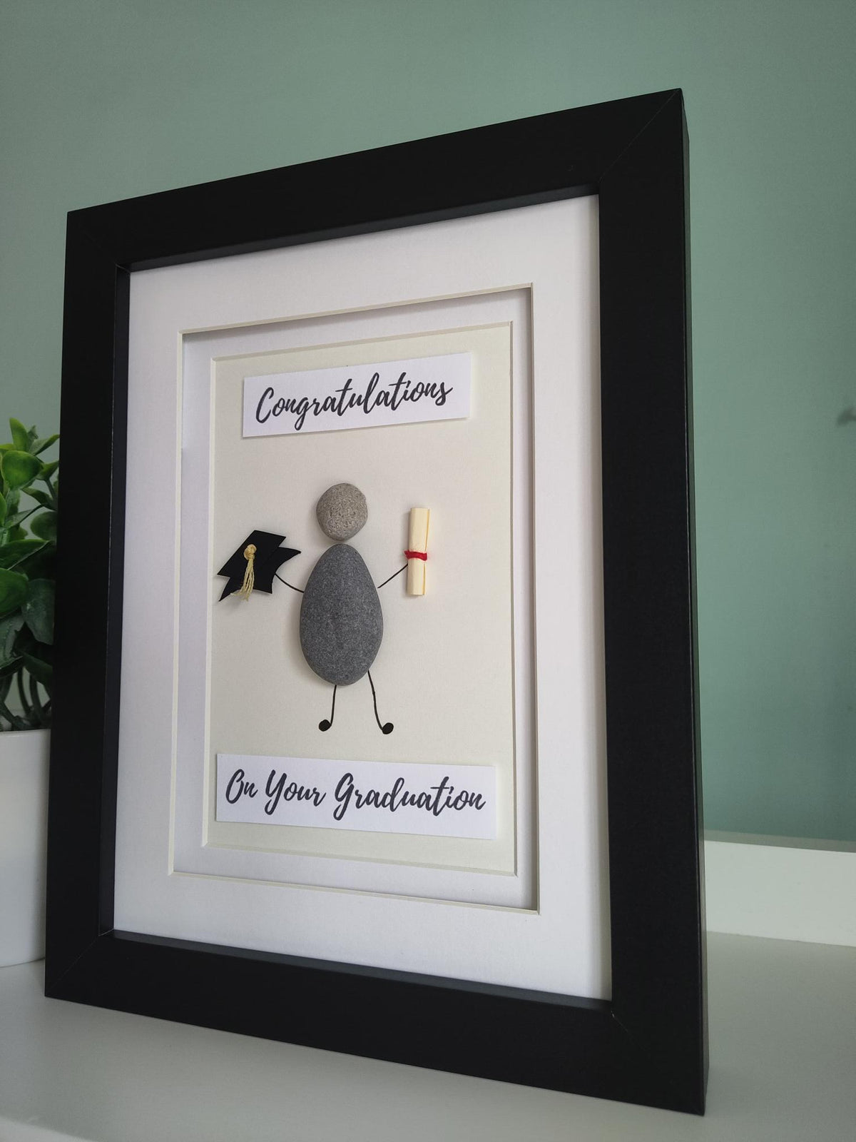 A side angle of a framed pebble art gift with the title 'CONGRATULATIONS ON YOUR GRADUATION' displayed, alongside a representation of a graduation cap and a graduation scroll.