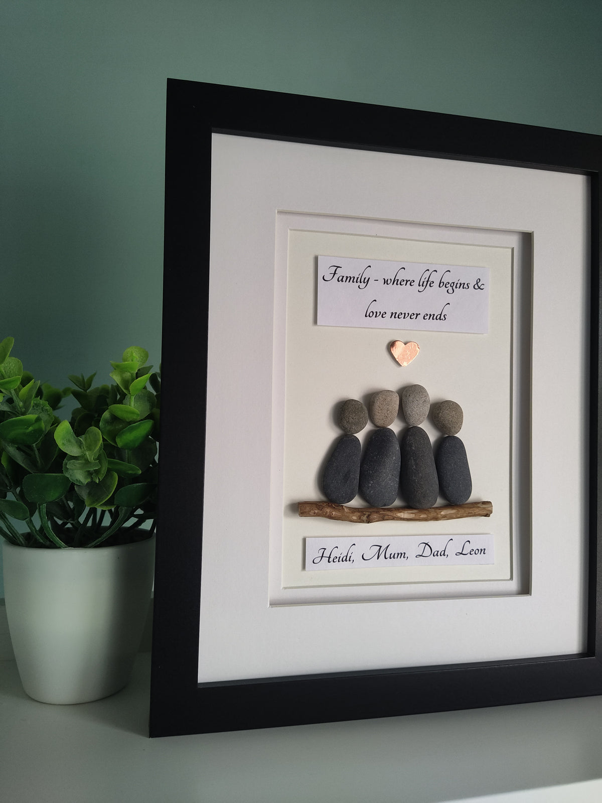 A side angle of a framed personalised family portrait featuring pebbles arranged in the shape of a family unit sitting on driftwood with personalised names, set against a white background in a black frame.