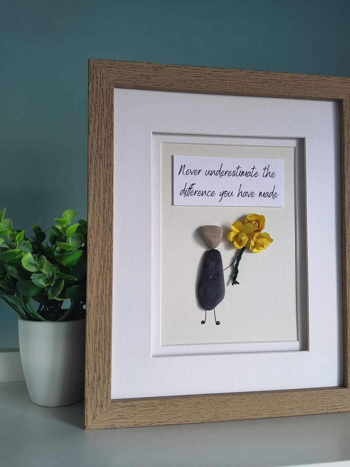A side angle of a framed pebble art gift featuring a figure holding a yellow bouquet of flowers and a quote stating 'Never Underestimate The Difference You Have Made' within a white mount and wooden design frame