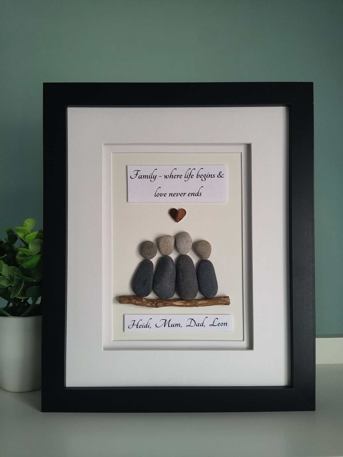 A framed personalised family portrait featuring pebbles arranged in the shape of a family unit sitting on driftwood with personalised names, set against a white background in a black frame.