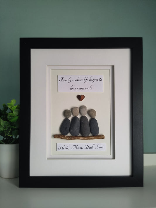 A framed personalised family portrait featuring pebbles arranged in the shape of a family unit sitting on driftwood with personalised names, set against a white background in a black frame.