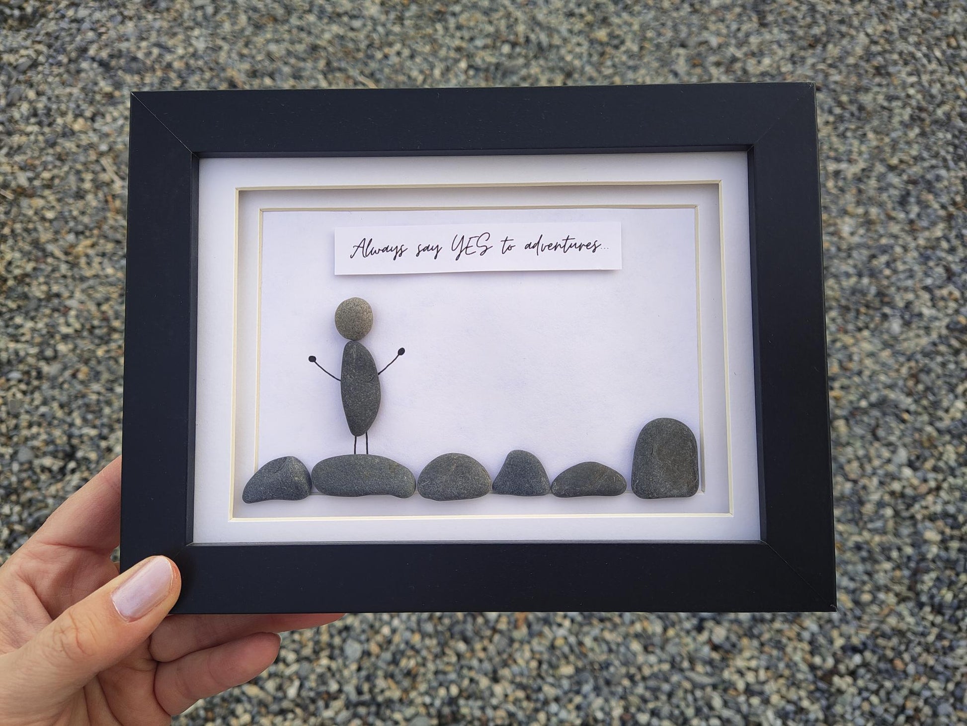 A hand holds a black-framed shadow box featuring a pebble figure and the phrase 'Always say YES to adventures' on a white background, set against a textured gray pebble surface.