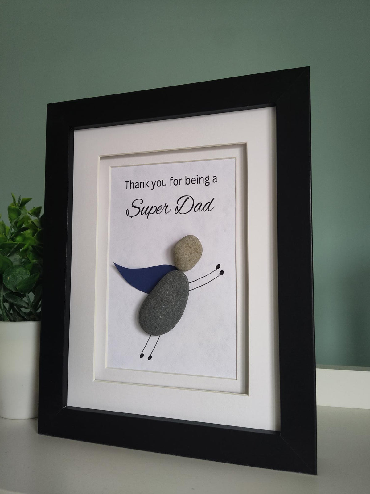 A side angle of a framed pebble art gift for 'Super Dad' with the message 'Thank you for being a Super Dad' displayed inside the frame, with a super hero theme pebble art character representing the super dad in a black frame 