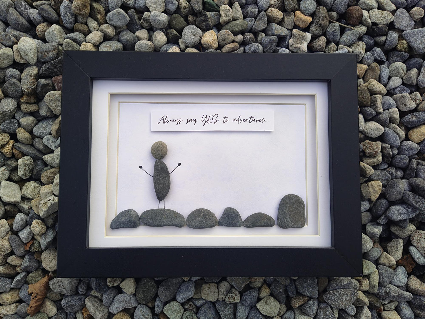 A framed pebble art piece displayed on a bed of small gray river stones. The black frame contains a white mat with a small stone figure standing on rocks, and a white banner above that reads, 'Always say YES to adventures'.