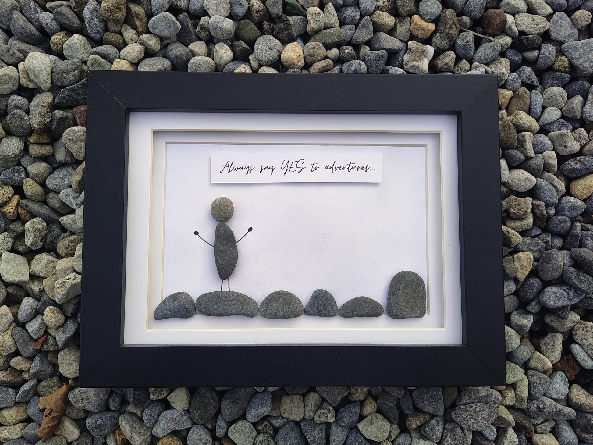 A framed pebble art piece displayed on a bed of small gray river stones. The black frame contains a white mat with a small stone figure standing on rocks, and a white banner above that reads, 'Always say YES to adventures'.