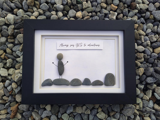 A framed pebble art piece displayed on a bed of small gray river stones. The black frame contains a white mat with a small stone figure standing on rocks, and a white banner above that reads, 'Always say YES to adventures'.