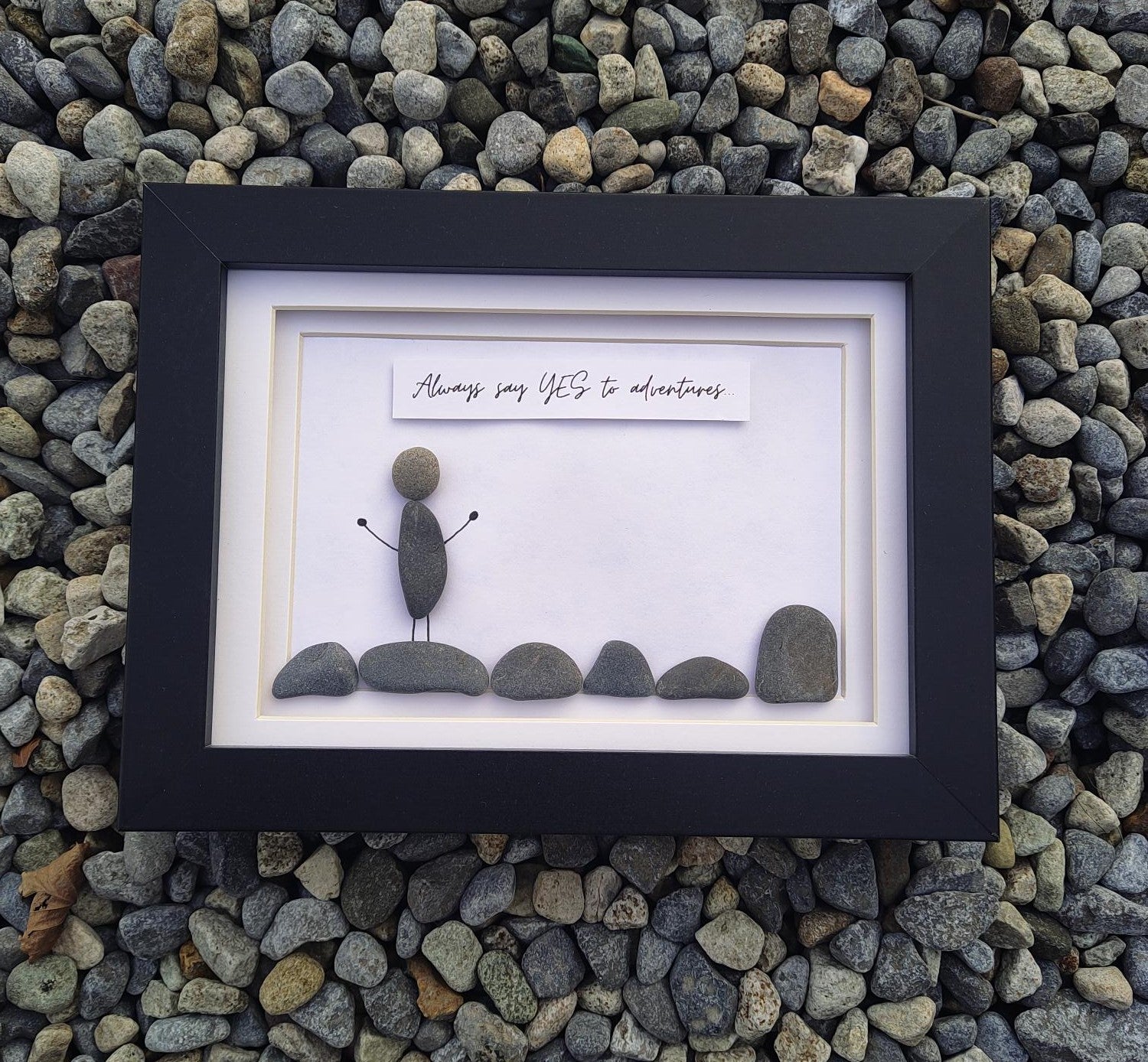A framed pebble art piece mounted on a bed of small gray river stones. The black frame contains a white mat with a small hand-drawn figure made of stones and a banner reading 'Always say YES to adventures'.