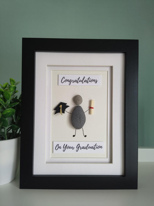A framed pebble art gift with the title 'CONGRATULATIONS ON YOUR GRADUATION' displayed, alongside a representation of a graduation cap and a graduation scroll.
