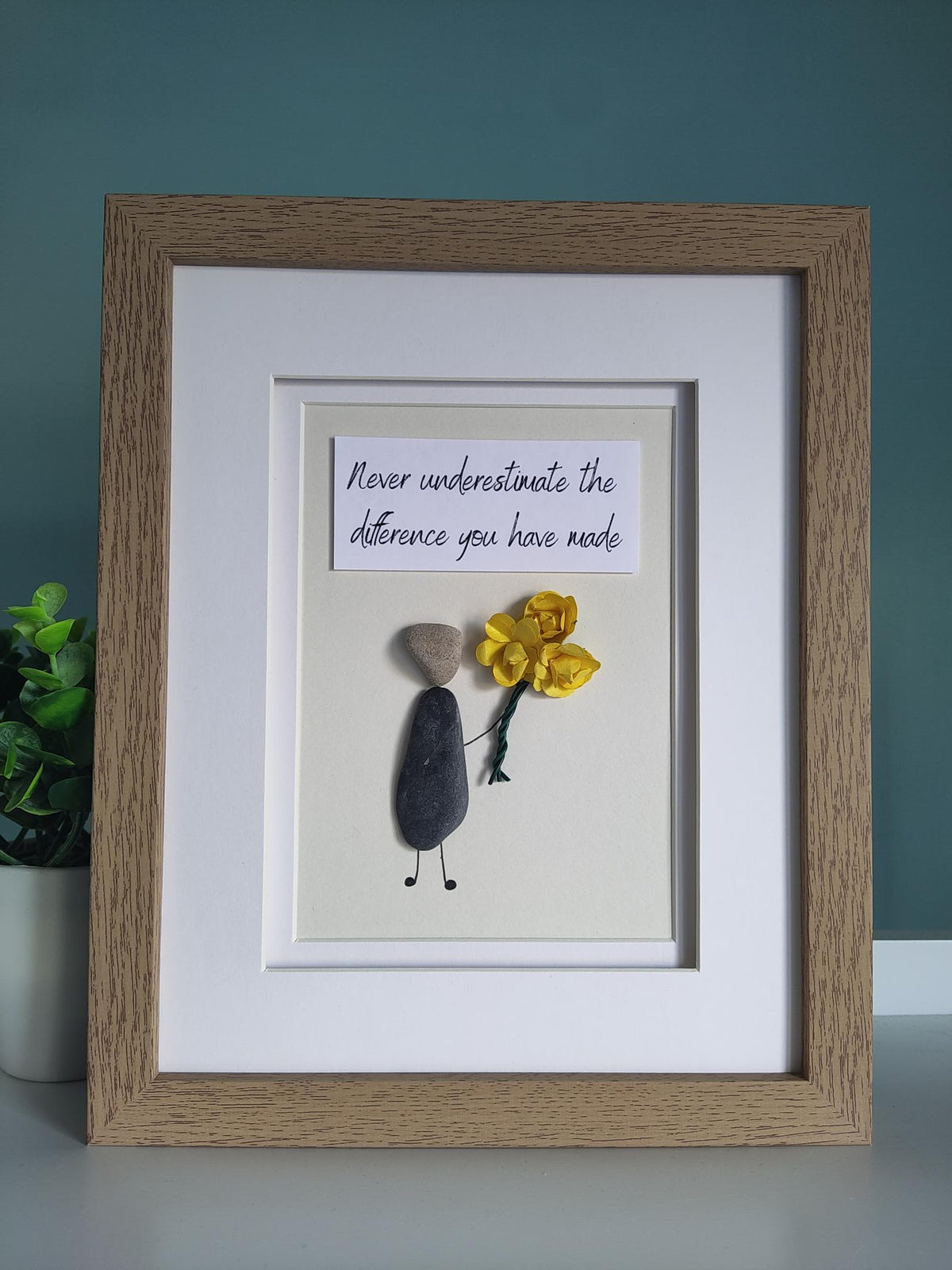 A framed pebble art gift featuring a figure holding a yellow bouquet of flowers and a quote stating 'Never Underestimate The Difference You Have Made' within a white mount and wooden design frame