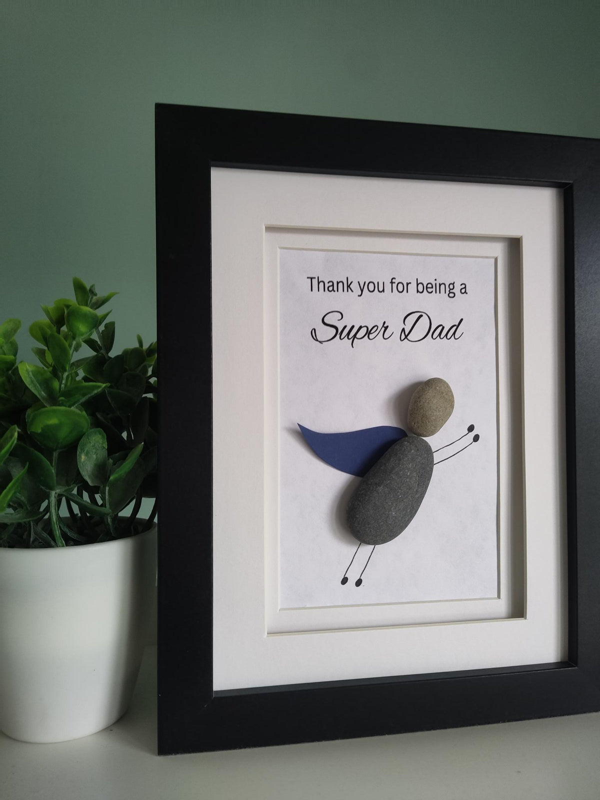 A side view of framed pebble art gift for 'Super Dad' with the message 'Thank you for being a Super Dad' displayed inside the frame, with a super hero theme pebble art character representing the super dad in a black frame 