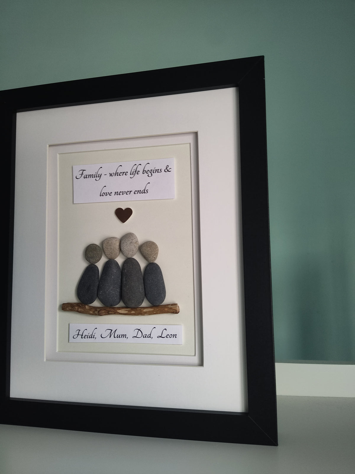 A side angle of a framed personalised family portrait featuring pebbles arranged in the shape of a family unit sitting on driftwood with personalised names, set against a white background in a black frame.