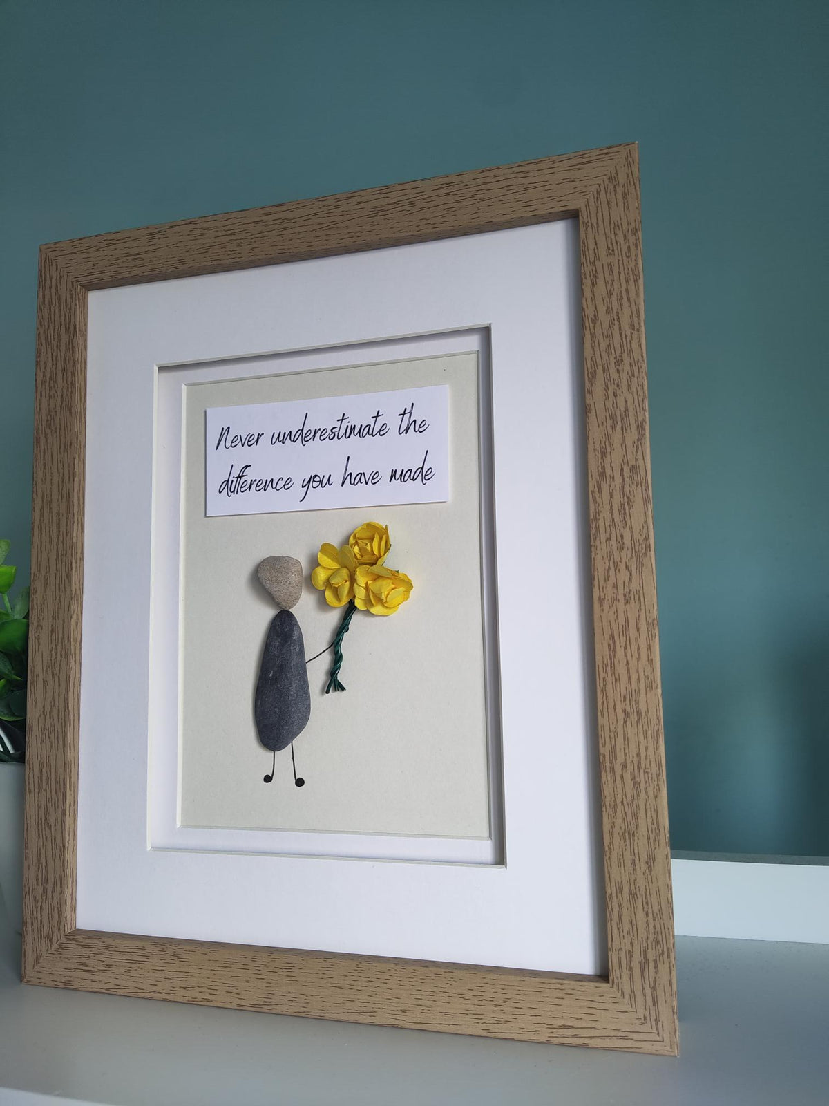 A side angle of a framed pebble art gift featuring a figure holding a yellow bouquet of flowers and a quote stating 'Never Underestimate The Difference You Have Made' within a white mount and wooden design frame