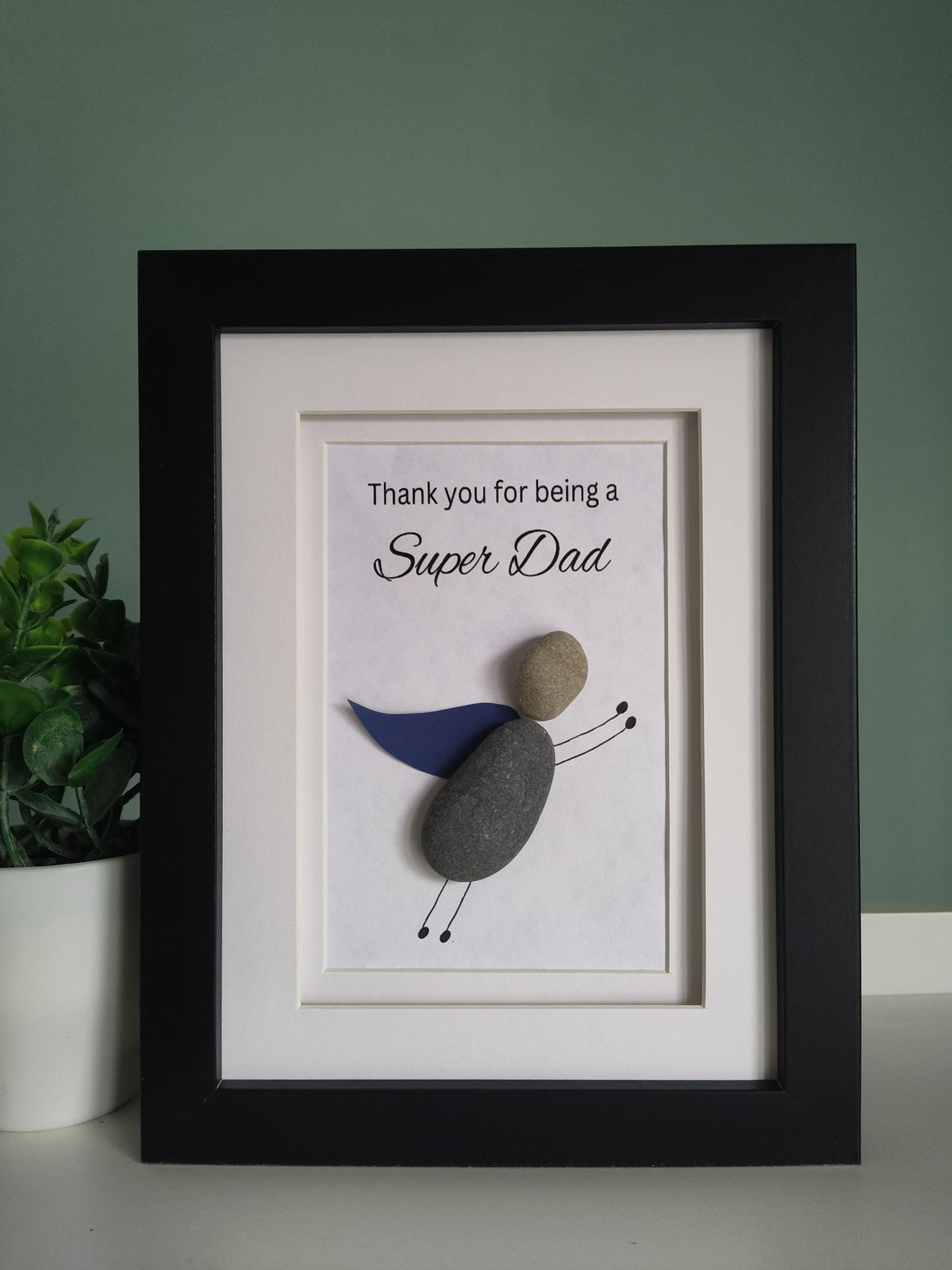 A framed pebble art gift for 'Super Dad' with the message 'Thank you for being a Super Dad' displayed inside the frame, with a super hero theme pebble art character representing the super dad in a black frame 